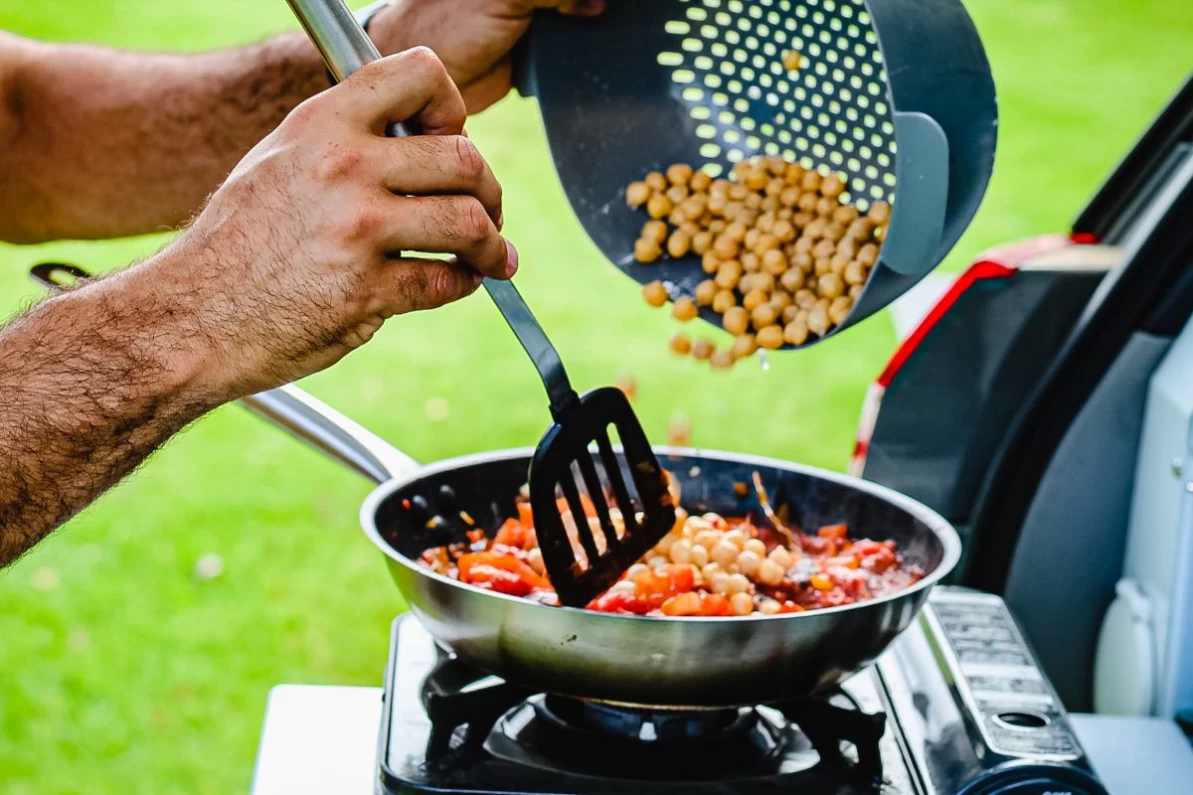 Close-up photo of a pan on the campig stove while the cook is adding chickpeas to the veggie stir fry in the pan